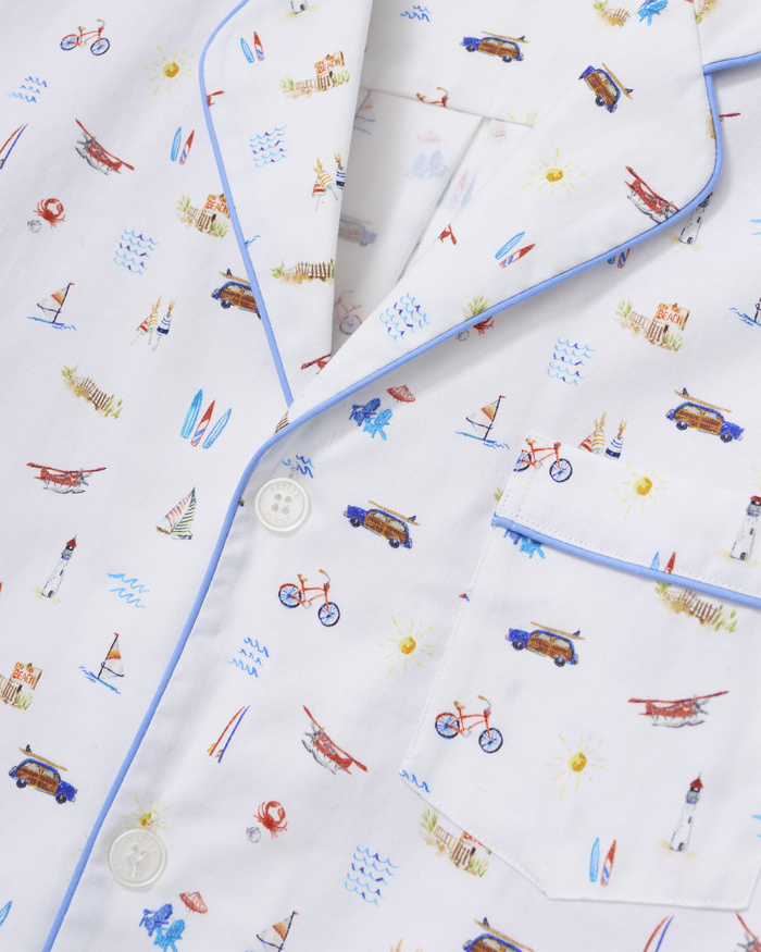 Men’s Twill Pajama Short Set in Summer Nostalgia features a white pajama top with blue trim, adorned with vibrant beach-themed illustrations such as cars, planes, bicycles, surfboards, lighthouses, suns, and sailboats. The shirt is finished with white buttons and a breast pocket.
