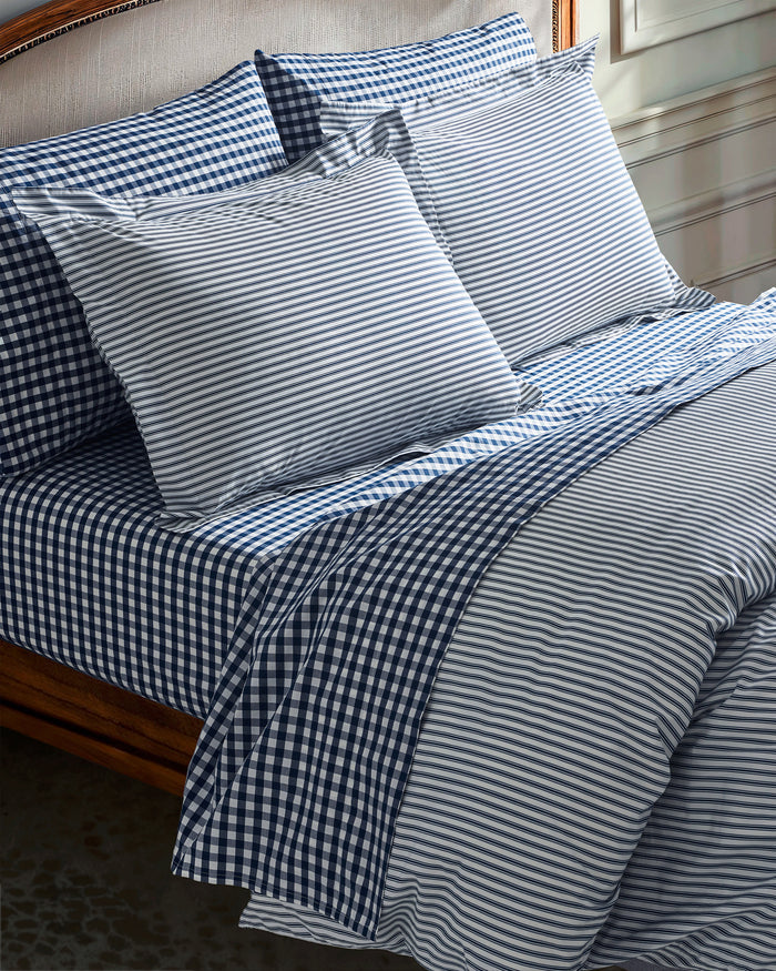 A neatly made bed featuring the Navy French Ticking and Navy Gingham Bedding Set on a wooden frame, with sunlight streaming onto the wall behind.
