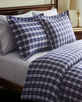 The Oxford Tartan Duvet and Navy French Ticking Bedding Set features navy blue and white plaid pillows and comforter, with striped white and gray bedsheets, next to a wooden nightstand topped with a lamp.