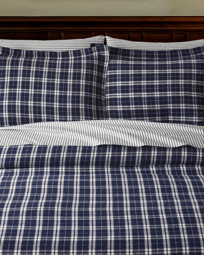 The Oxford Tartan Duvet and Navy French Ticking Bedding Set features navy blue and white plaid pillows and comforter, with striped white and gray bedsheets, next to a wooden nightstand topped with a lamp.