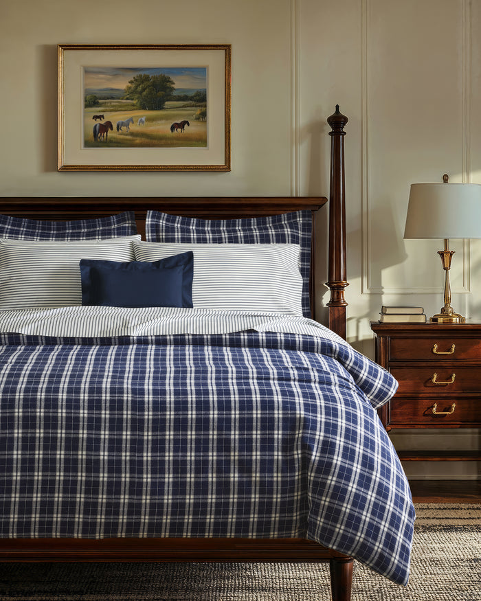 The Oxford Tartan Duvet and Navy French Ticking Bedding Set features navy blue and white plaid pillows and comforter, with striped white and gray bedsheets, next to a wooden nightstand topped with a lamp.