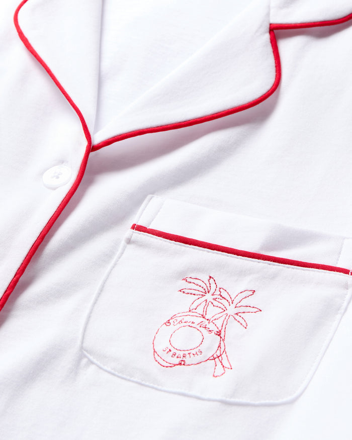 Close-up of the Petite Plume x Eden Rock St Barth Mens White Pima Pajama Set. This limited-edition top features red piping on the collar, lapel, and pocket edges, with a red embroidered lifebuoy and palm trees design saying Ocean Dream and YEARTIS, capturing Caribbean paradise allure.