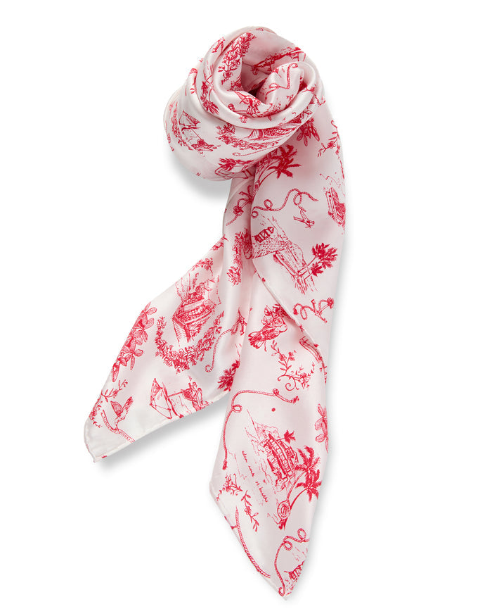 The Petite Plume x Eden Rock Reverie Silk Scarf is a white silk accessory with red toile-style illustrations, featuring intricate scenes and floral motifs, loosely rolled at the top and elegantly draped.