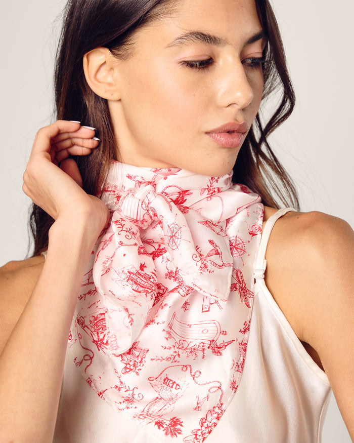The Petite Plume x Eden Rock Reverie Silk Scarf is a white silk accessory with red toile-style illustrations, featuring intricate scenes and floral motifs, loosely rolled at the top and elegantly draped.