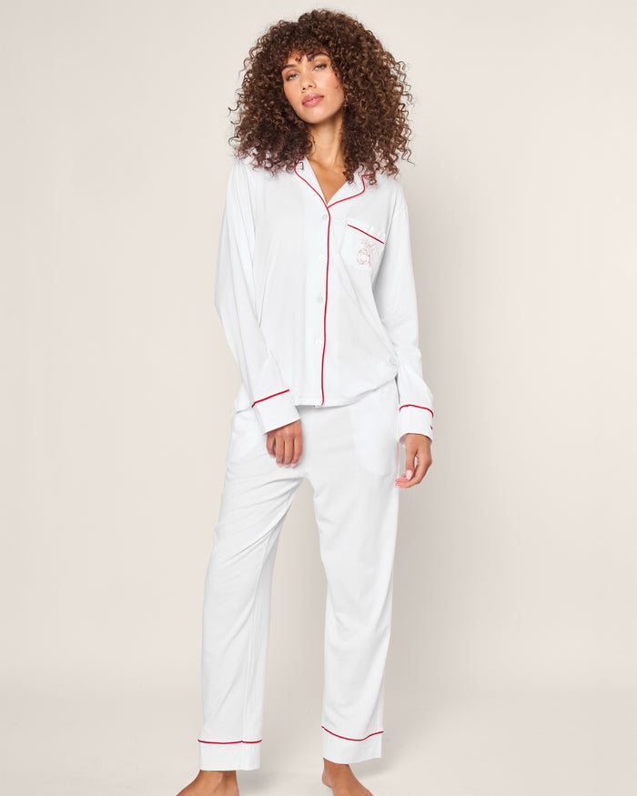 A person with curly hair wears a luxury sleepwear set from Petite Plume—the Womens White Pima Pajama Set with red piping from their limited-edition collection. The button-up top and long pants pop against a plain, light-colored background as they face the camera.