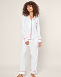 A person with curly hair wears a luxury sleepwear set from Petite Plume—the Womens White Pima Pajama Set with red piping from their limited-edition collection. The button-up top and long pants pop against a plain, light-colored background as they face the camera.