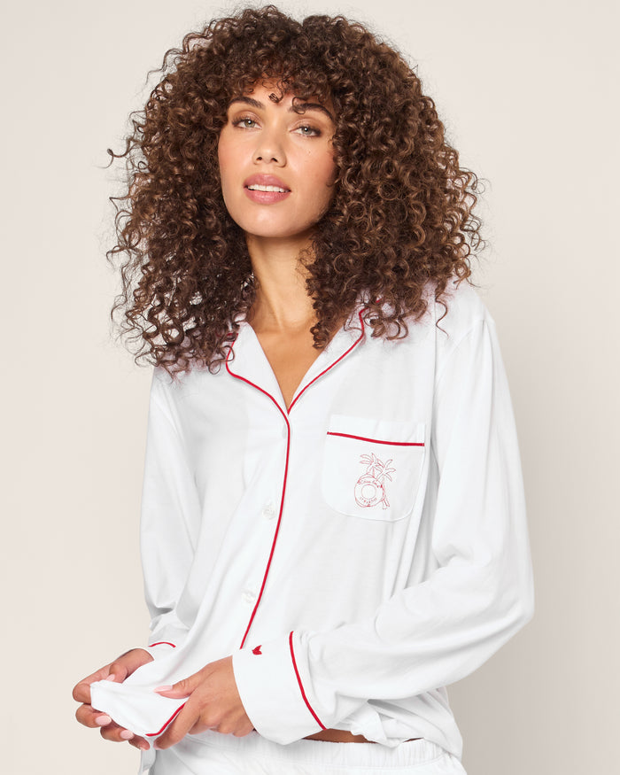 A person with curly hair models the Petite Plume x Eden Rock St Barth Womens White Pima Pajama top, featuring red piping and a small embroidered pocket design from this limited-edition luxury sleepwear collection, against a plain background, gently holding its hem.