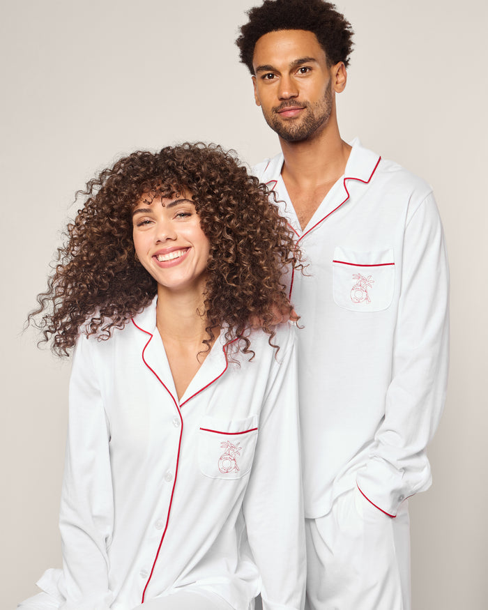 Two people in matching Petite Plume x Eden Rock St Barth Womens White Pima Pajama Sets, featuring red piping and an elegant embroidered pocket design, stand against a Caribbean-inspired backdrop; the seated one smiles, curly-haired, while the other stands calmly with short hair.
