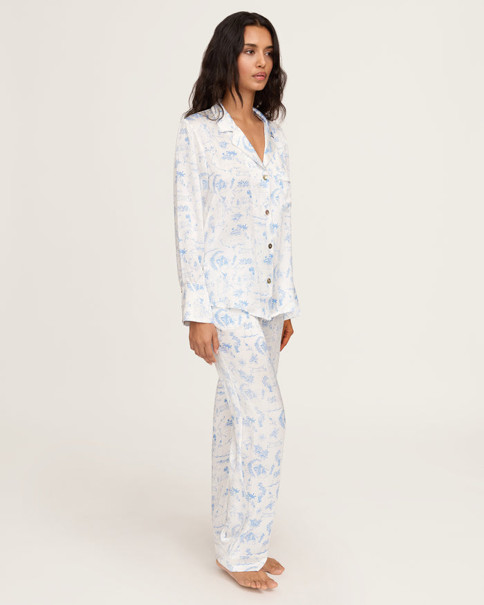 The Petite Plume x Eden Rock Blue Reverie Women's Silk Pajama Set features white pajamas with a blue floral and animal print, a long-sleeve collared button-up top, and matching pants with an elastic waistband.