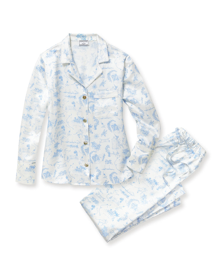 The Petite Plume x Eden Rock Blue Reverie Women's Silk Pajama Set features white pajamas with a blue floral and animal print, a long-sleeve collared button-up top, and matching pants with an elastic waistband.