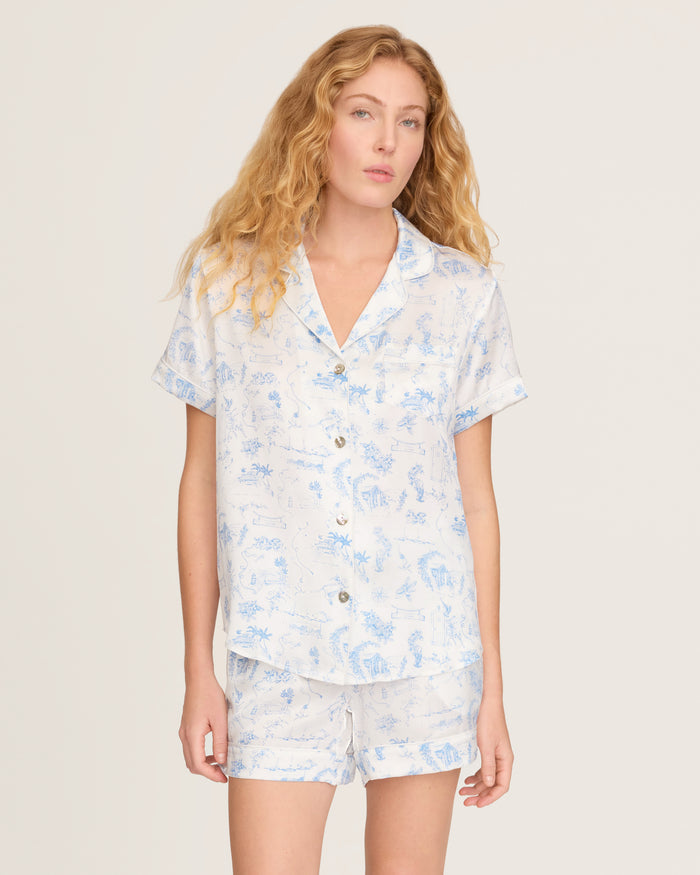 The Petite Plume x Eden Rock Blue Reverie Women's Silk Short Set features a white base with light blue nature-inspired prints, a short-sleeve button-up shirt with chest pocket, and drawstring waist shorts.
