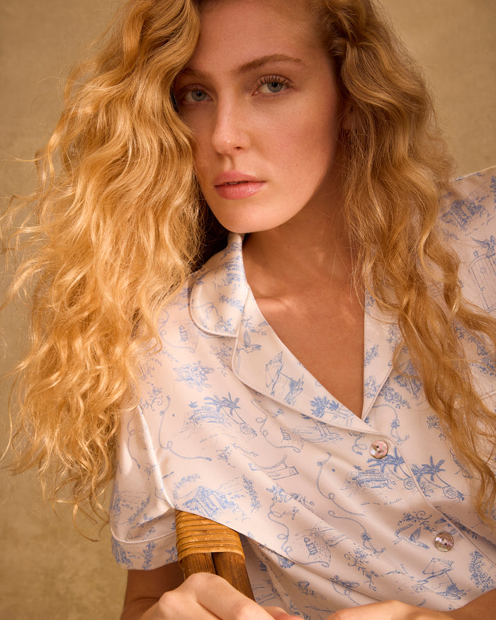 The Petite Plume x Eden Rock Blue Reverie Women's Silk Short Set features a white base with light blue nature-inspired prints, a short-sleeve button-up shirt with chest pocket, and drawstring waist shorts.