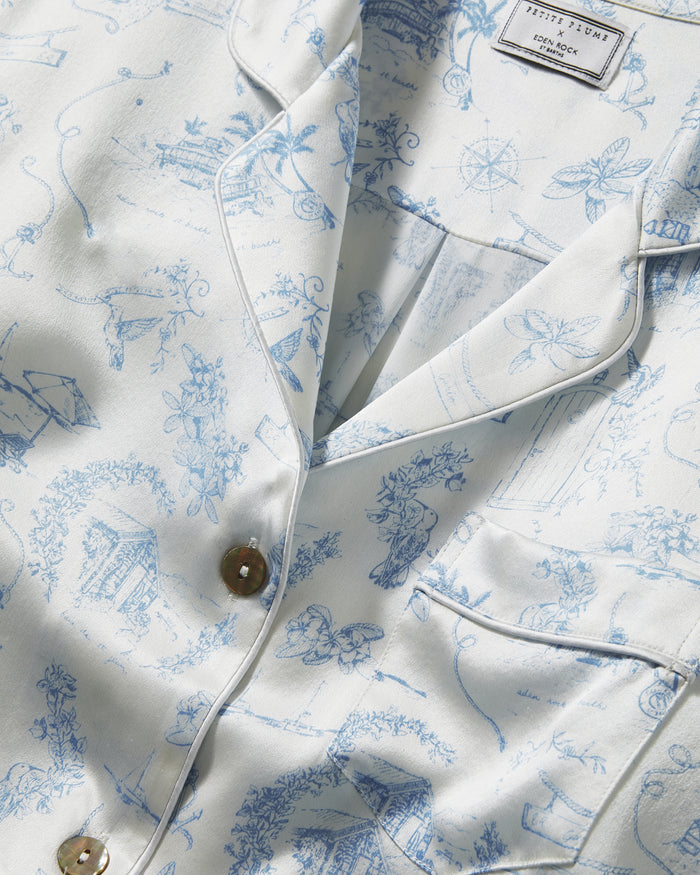 The Petite Plume x Eden Rock Blue Reverie Women's Silk Short Set features a white base with light blue nature-inspired prints, a short-sleeve button-up shirt with chest pocket, and drawstring waist shorts.