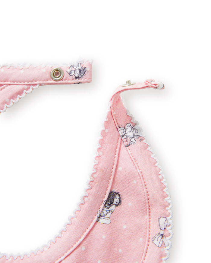 The Petite Plume x Eloise Baby Pima Bib in Eloise Adventures features pink fabric with white polka dots, playful black-and-white drawings, whimsical motifs, a scalloped edge, and a convenient snap closure at the neck.