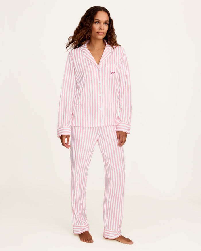 The Petite Plume x Eloise Women's Pima Pajama Set in Eloise Classic Stripe features pink and white vertical stripes, a long-sleeve button-up top with a collar, chest pocket with red embroidery, and matching pants.