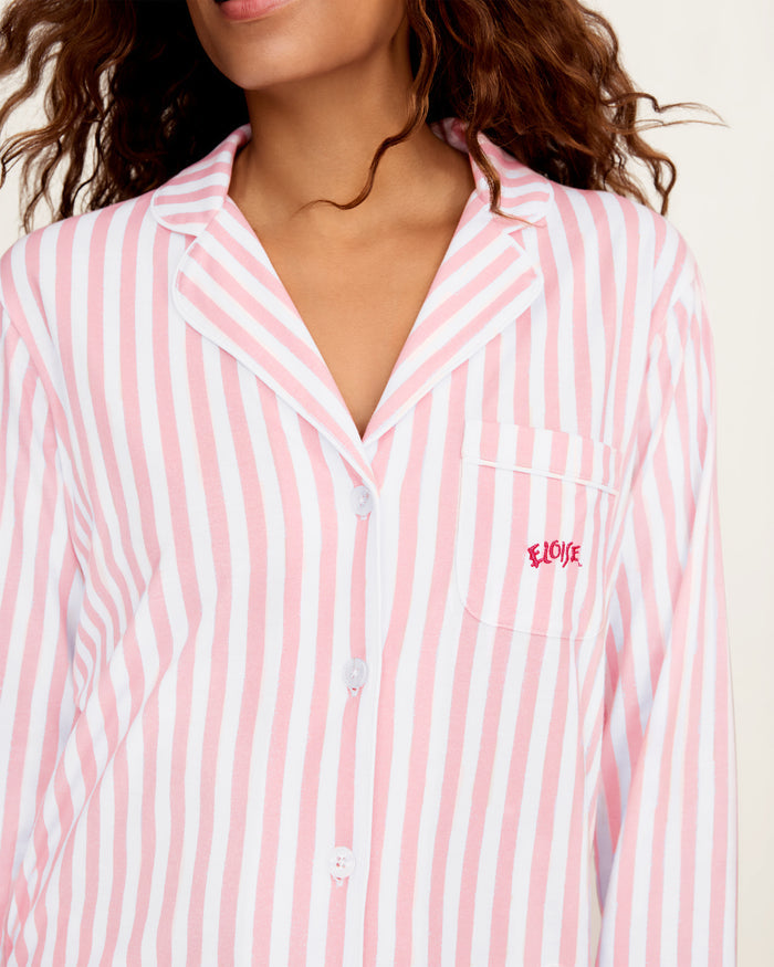 The Petite Plume x Eloise Women's Pima Pajama Set in Eloise Classic Stripe features pink and white vertical stripes, a long-sleeve button-up top with a collar, chest pocket with red embroidery, and matching pants.