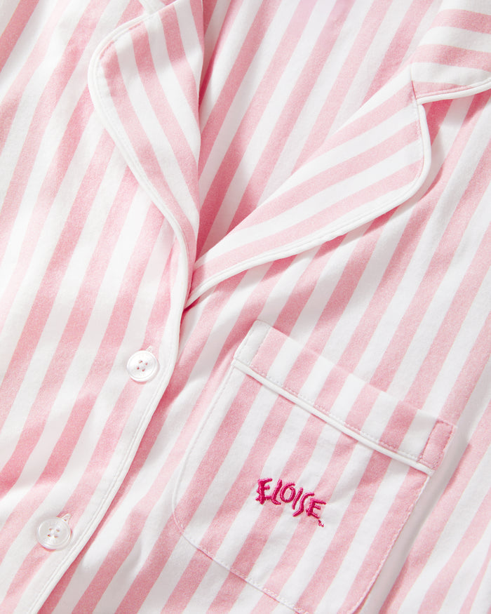 The Petite Plume x Eloise Women's Pima Pajama Set in Eloise Classic Stripe features pink and white vertical stripes, a long-sleeve button-up top with a collar, chest pocket with red embroidery, and matching pants.