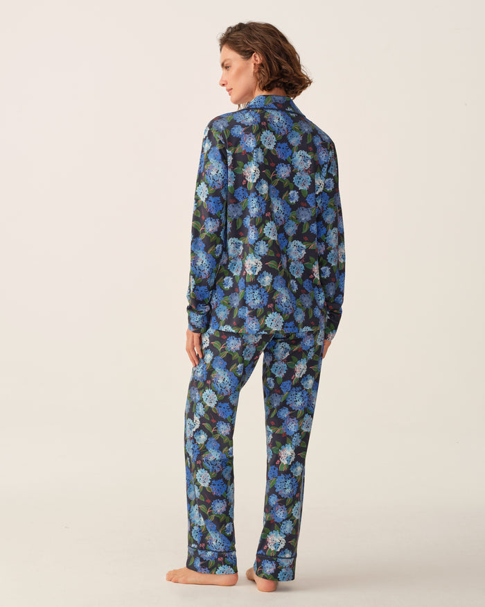 A person stands barefoot, facing away, wearing the Petite Plume x Hedges Women’s Pima Pajama Set in Hydrangea—featuring a blue and purple floral pattern on a dark background—against a plain, light backdrop.