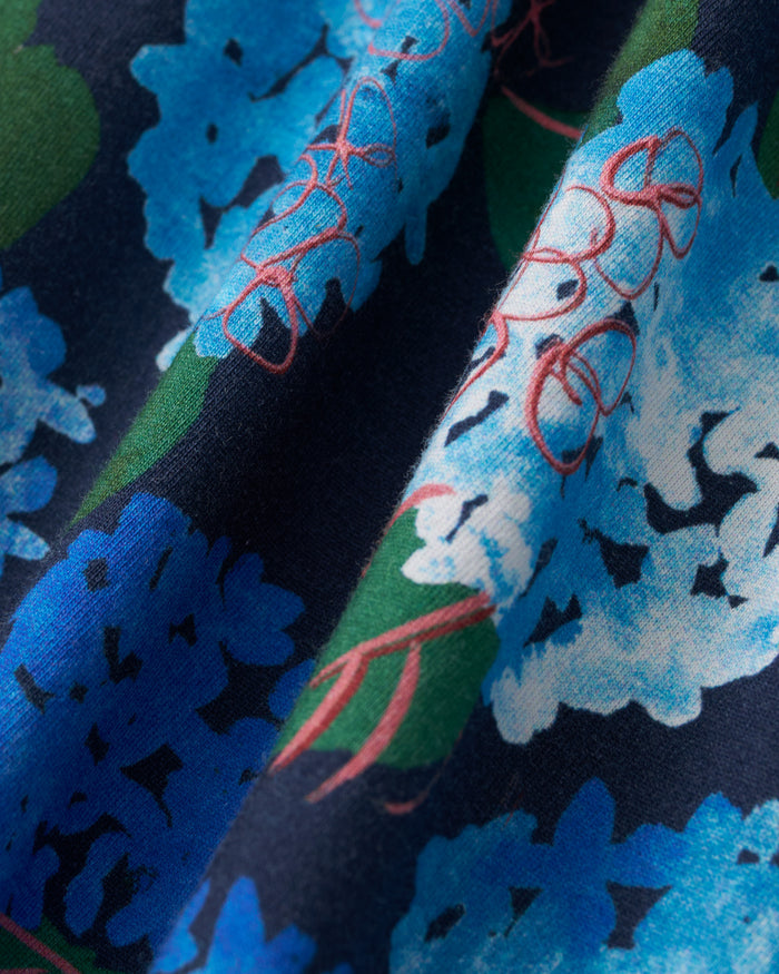 Close-up of the Petite Plume x Hedges Women’s Pima Pajama Set in Hydrangea, featuring fabric with a floral pattern in shades of blue and white, set against a dark blue background, accented with green leaves and pink flower outlines. The soft pima cotton drapes in gentle folds.