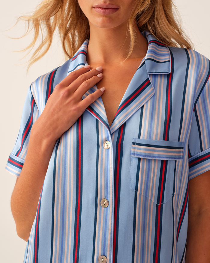 A woman models the Petite Plume x Hedges Women’s Silk Short Set in Blue Stripe—a light blue, short-sleeved, button-up shirt featuring vertical red, white, and navy stripes and a chest pocket. She gently touches her collar with one hand.