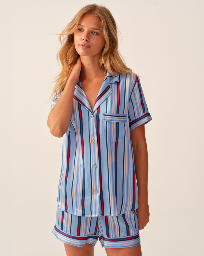 A person with long blonde hair wears the Petite Plume x Hedges Women’s Silk Short Set in Blue Stripe, featuring a vertically striped short-sleeve button-up top and matching shorts, standing against a plain light background.