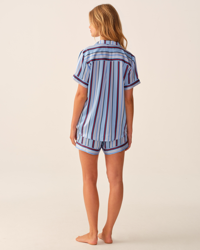A woman stands barefoot, facing away, wearing the Petite Plume x Hedges Women’s Silk Short Set in Blue Stripe—a short-sleeved, button-up shirt and matching shorts with blue, white, and red vertical stripes—against a light, plain background.
