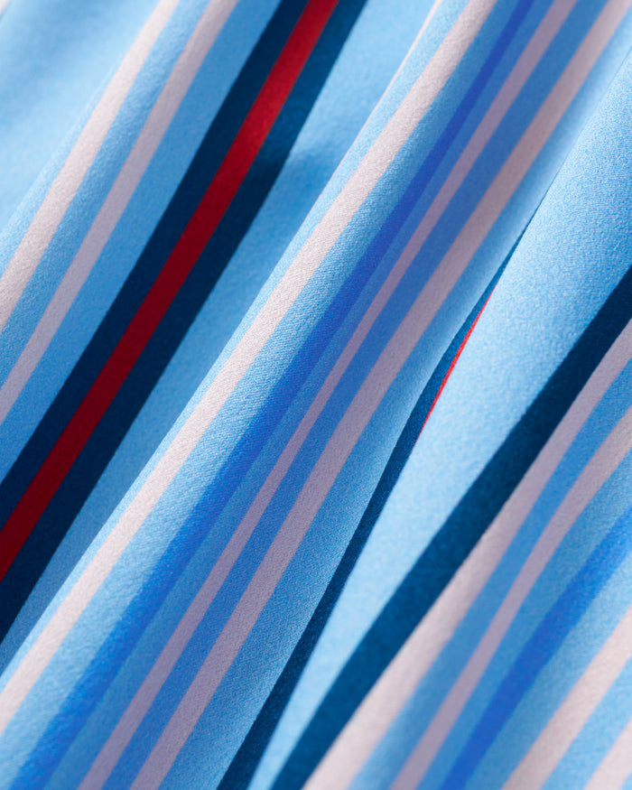 Close-up of the Petite Plume x Hedges Women’s Silk Short Set in Blue Stripe, featuring smooth blue silk fabric adorned with diagonal stripes in varying shades of blue, white, and red. Soft folds create subtle shadows on the slightly shiny surface.