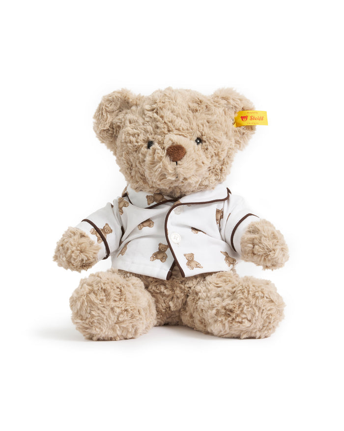 The Petite Plume x Steiff Teddy Bear, a plush light brown bear in a white shirt with teddy bear prints and a yellow ear tag, sits on a bed against a dark brown pillow.