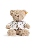 The Petite Plume x Steiff Teddy Bear, a plush light brown bear in a white shirt with teddy bear prints and a yellow ear tag, sits on a bed against a dark brown pillow.
