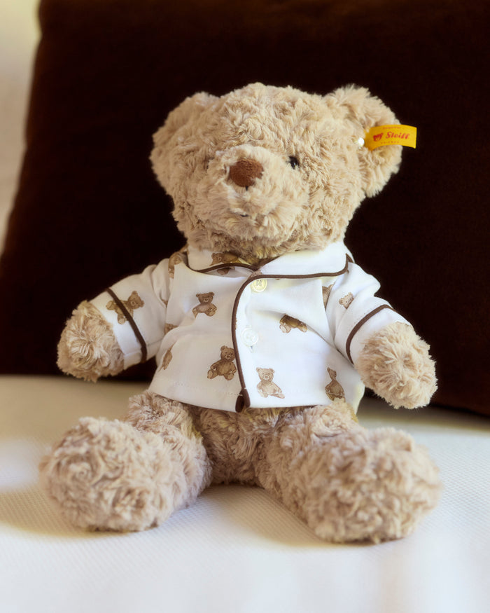 The Petite Plume x Steiff Teddy Bear, a plush light brown bear in a white shirt with teddy bear prints and a yellow ear tag, sits on a bed against a dark brown pillow.