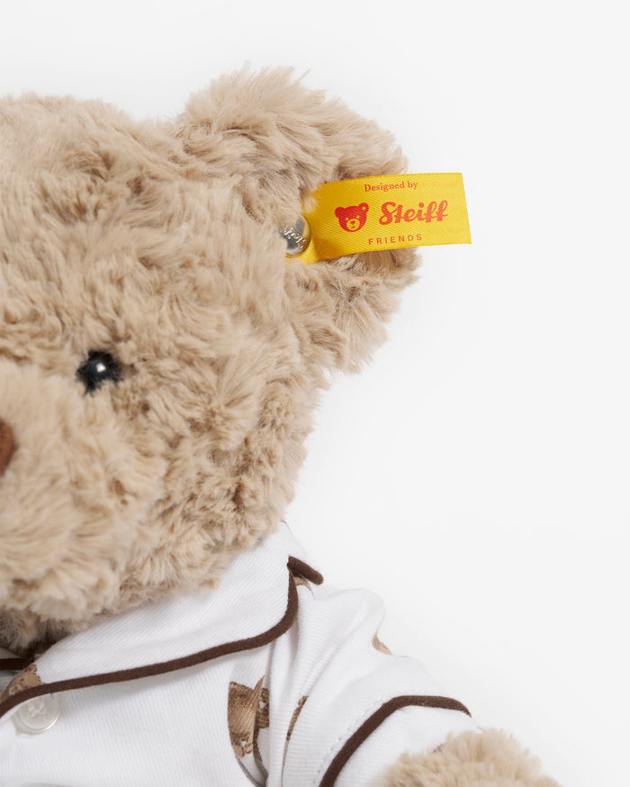 The Petite Plume x Steiff Teddy Bear, a plush light brown bear in a white shirt with teddy bear prints and a yellow ear tag, sits on a bed against a dark brown pillow.