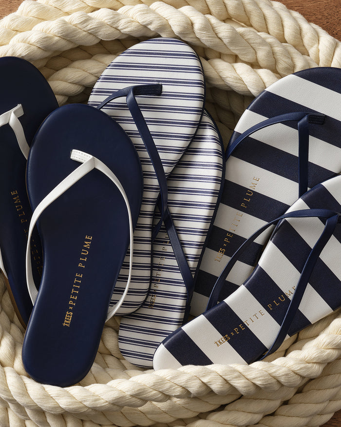 A basket filled with three pairs of Petite Plume x TKEES Lily Sandal in Navy French Ticking: one in solid navy, one featuring navy and white stripes, and one showcasing navy and white horizontal stripes, each adorned with MES PETITE PLUME written in gold on the soles.