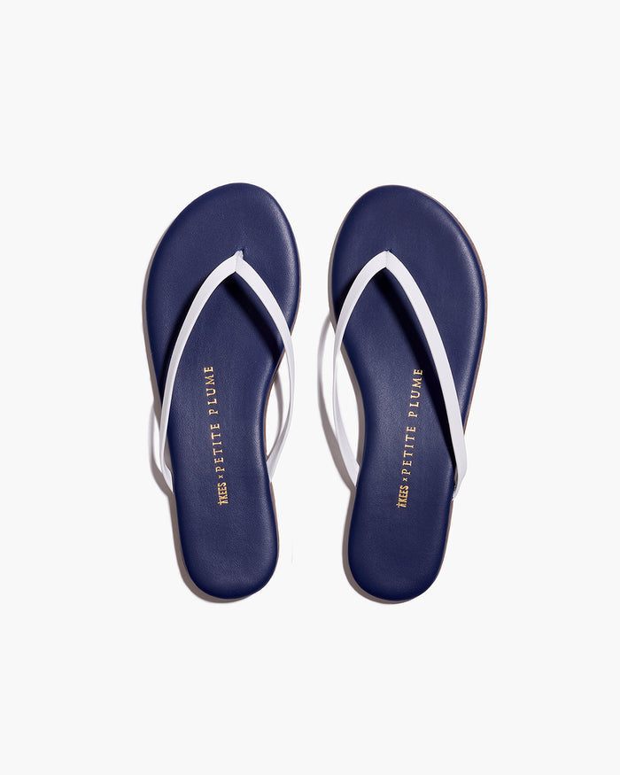 A pair of Petite Plume x TKEES Lily Sandals in navy with white straps, photographed from above on a white background. "THE PETITE PLUME" is printed in gold on the insoles.