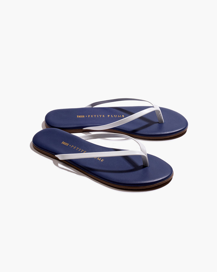 A pair of Petite Plume x TKEES Lily Sandals in navy with white straps, photographed from above on a white background. "THE PETITE PLUME" is printed in gold on the insoles.