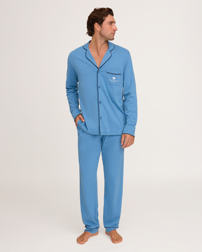 A man stands barefoot on a white background, looking to his right, wearing the Petite Plume x White Elephant Men’s Pima Pajama Set in blue—a long-sleeve, button-up top with matching pants.