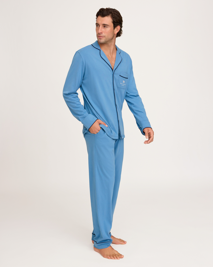 A person stands barefoot on a white background, wearing the Petite Plume x White Elephant Men’s Pima Pajama Set in light blue with dark trim and a chest pocket. The pajama set features a button-up top and matching pants. The person is looking slightly to the side.