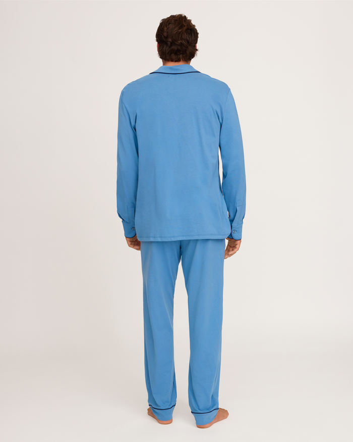 A person stands barefoot facing away from the camera, wearing the Petite Plume x White Elephant Men’s Pima Pajama Set in light blue, featuring a matching long-sleeve top and pants, against a plain white background.
