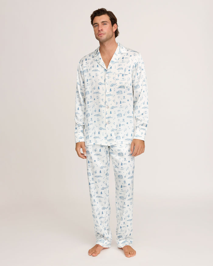A man stands barefoot against a plain white background, wearing the Petite Plume x White Elephant Men’s Silk Pajama Set—a matching ensemble of white pajama pants and a long-sleeve button-up shirt featuring a subtle light blue pattern.