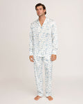 A man stands barefoot against a plain white background, wearing the Petite Plume x White Elephant Men’s Silk Pajama Set—a matching ensemble of white pajama pants and a long-sleeve button-up shirt featuring a subtle light blue pattern.