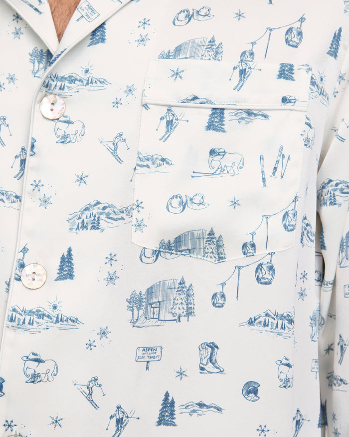 Close-up of the Petite Plume x White Elephant Men’s Silk Pajama Set in white, featuring blue line drawings of winter and mountain scenes—including skiers, trees, cabins, gondolas, and animals—with a visible chest pocket and buttons.