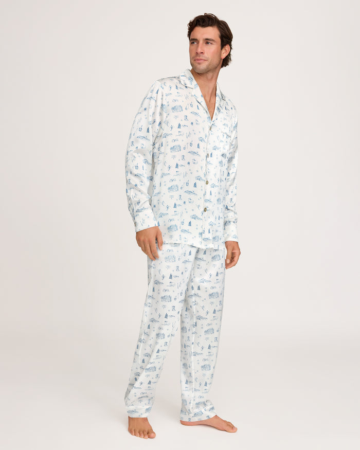 A man stands barefoot, wearing the Petite Plume x White Elephant Men’s Silk Pajama Set—white pajama pants and a long-sleeve button-up shirt with a light blue pattern—against a plain light background.