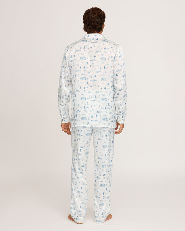 A person stands barefoot facing away, wearing the Petite Plume x White Elephant Men’s Silk Pajama Set, which features a subtle blue pattern on light-colored fabric. The background is plain and white.