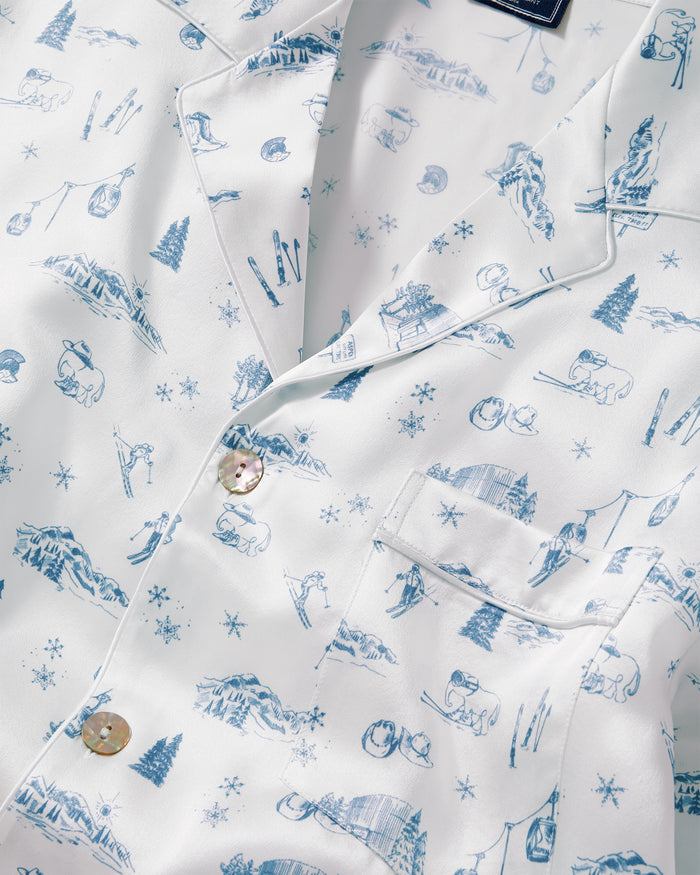 Close-up of the Petite Plume x White Elephant Men’s Silk Pajama Set, featuring a white button-up shirt adorned with blue winter-themed illustrations—mountains, skis, trees, snowflakes, cabins, and people skiing—scattered across the luxurious silk fabric.