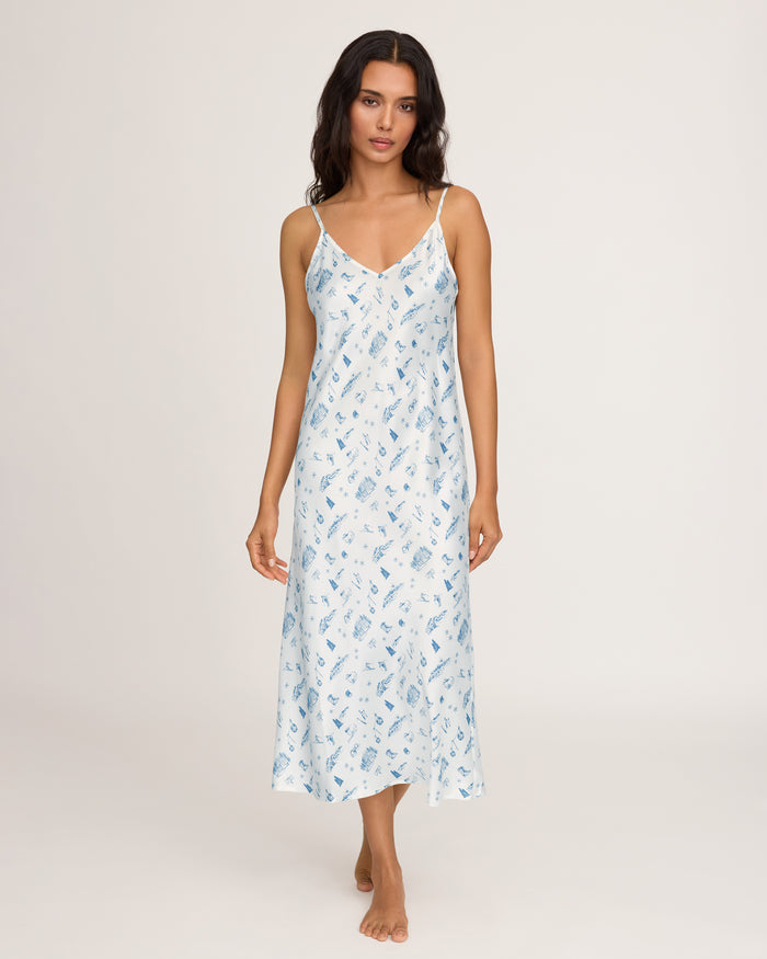 A woman stands barefoot on a plain background, facing forward with a relaxed expression, wearing the Petite Plume x White Elephant Women’s Silk Cosette Nightgown—an ankle-length white slip dress adorned with blue floral patterns.