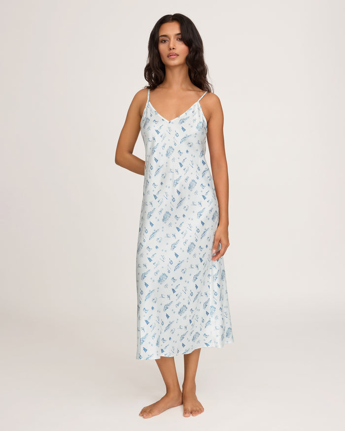 A woman stands barefoot against a plain background, wearing the Petite Plume x White Elephant Women’s Silk Cosette Nightgown—a light blue, sleeveless, V-neck, ankle-length gown adorned with a delicate blue pattern. Her dark hair is loose and she has a neutral expression.