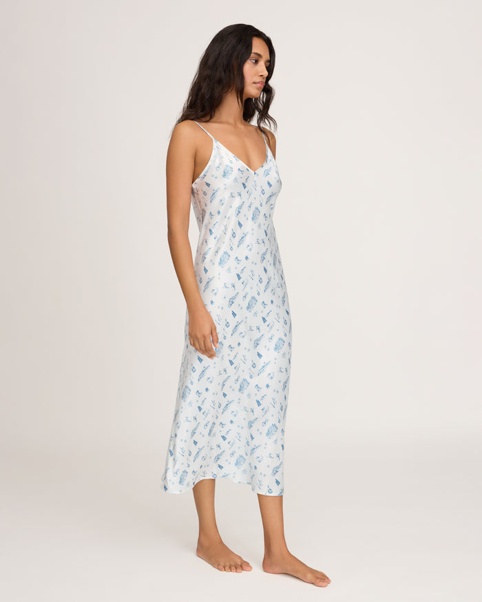 A woman stands barefoot against a plain white background, wearing the Petite Plume x White Elephant Women’s Silk Cosette Nightgown—a light blue, sleeveless, ankle-length gown with thin straps and a subtle printed pattern.