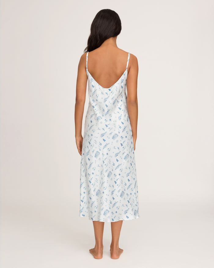 A woman with long dark hair stands barefoot, facing away from the camera, wearing the Petite Plume x White Elephant Women’s Silk Cosette Nightgown—a light blue, sleeveless, patterned gown with thin straps and a low back—set against a plain light background.