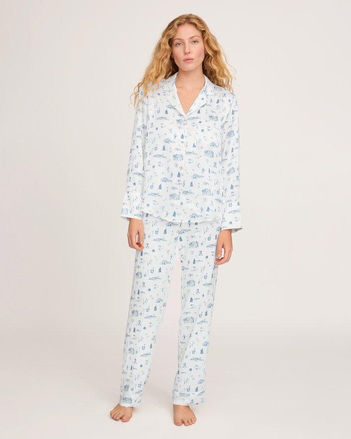 A woman with long, wavy blonde hair stands barefoot against a plain white background, wearing the Petite Plume x White Elephant Women’s Silk Pajama Set—a light blue and white printed button-up shirt and pants.