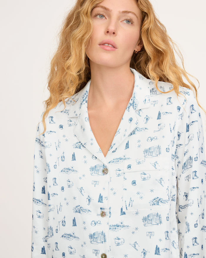 A woman with long wavy blonde hair wears the Petite Plume x White Elephant Women’s Silk Pajama Set—a white button-up top featuring blue winter-themed illustrations—while looking slightly upward with a neutral expression against a plain light background.