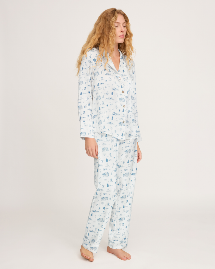 A woman with long wavy hair stands barefoot against a plain white background, wearing the Petite Plume x White Elephant Women’s Silk Pajama Set—a light blue and white printed pajama ensemble featuring a button-up top and matching pants.